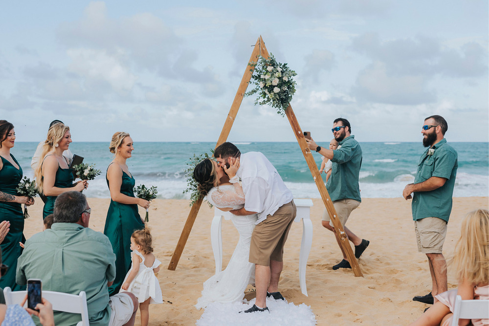 A bride and groom kiss on the beach during their wedding ceremony.