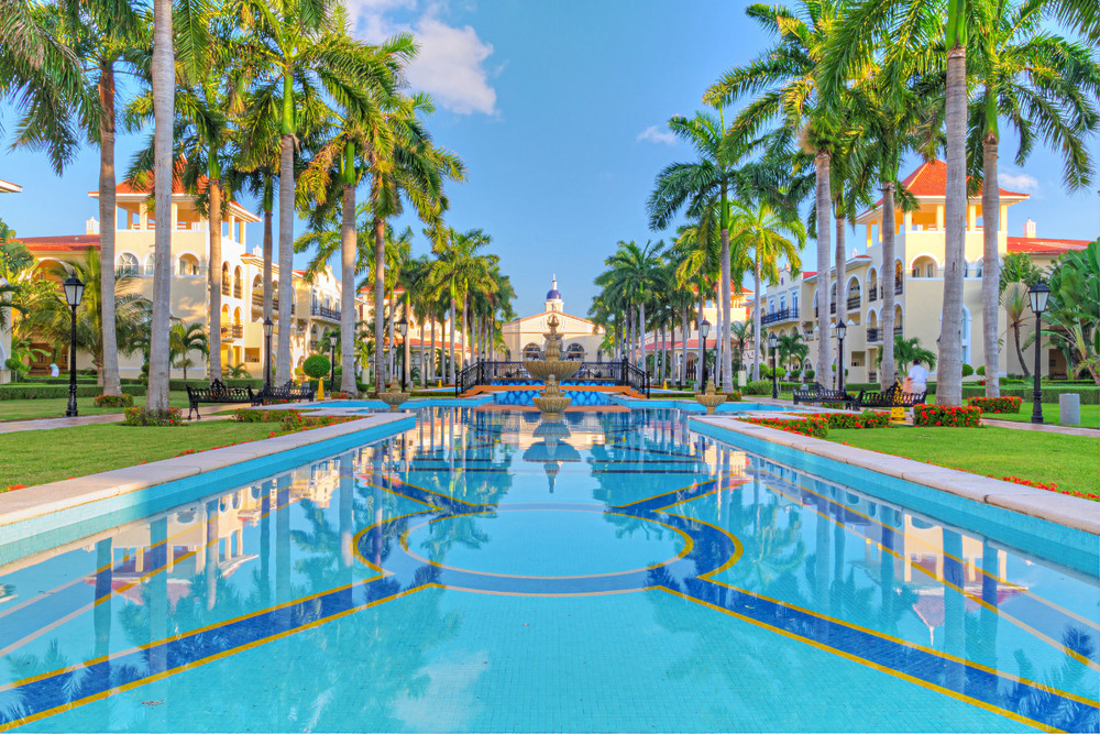 A pool surrounded by palm trees in front of a hotel, set up for a wedding.