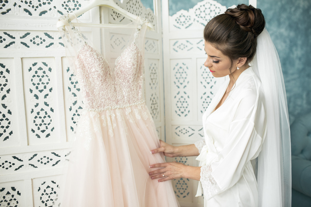 A bride in a white robe admires wedding dress trends, gazing at a lace gown for her destination wedding.