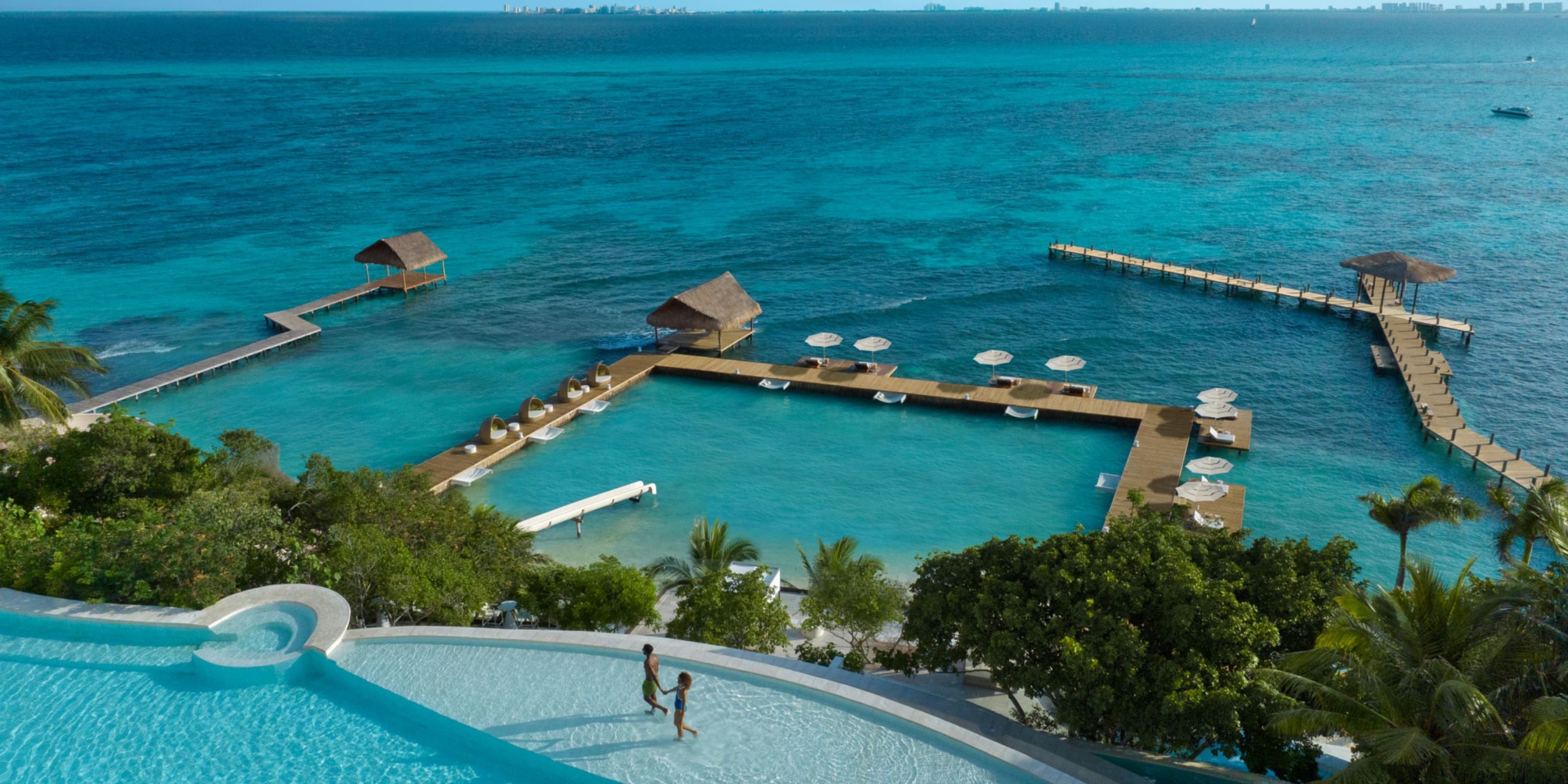Infinity pool at Impression Isla Mujeres by Secrets, offering a breathtaking view of the turquoise ocean with wooden docks and thatched structures stretching into the water, all enveloped by lush greenery.