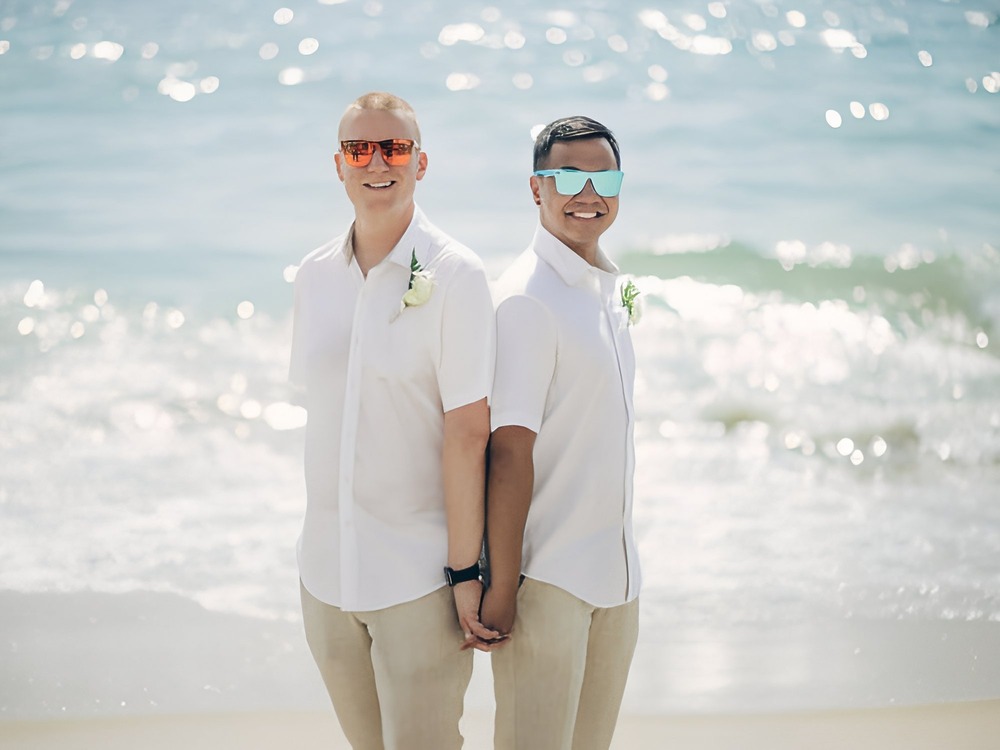 Two men in white shirts and sunglasses hold hands, dreaming about where to begin as they plan their destination wedding.