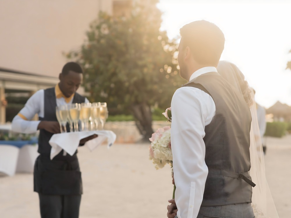 A waiter serves champagne to a couple as they discuss where to begin with their wedding plans, considering whether a destination wedding might be the perfect choice.