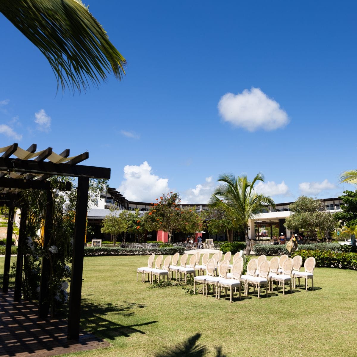 White chairs set up on a green lawn for a destination wedding, with palm trees and clear blue sky above.
