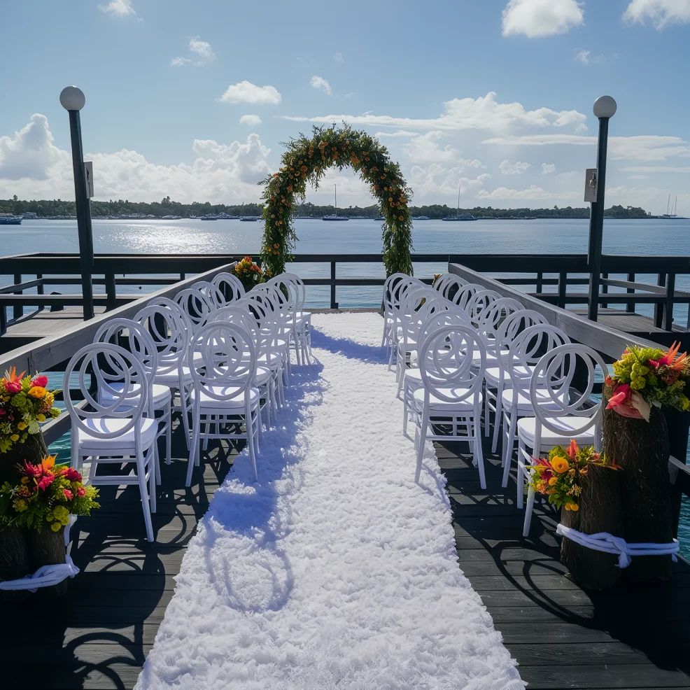 Outdoor wedding setup on a dock with white chairs, a floral arch, and a white carpet aisle overlooking a body of water.