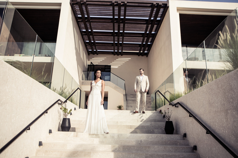 A bride in white and a groom in a suit stand separately on contemporary stairs, captured in an elegant destination wedding photography style.