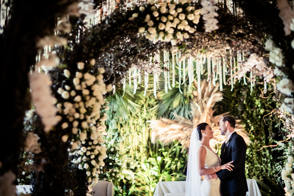 The bride and groom stand together under a floral arch, captured in timeless wedding photography style reminiscent of classic films—perfect for a destination wedding.