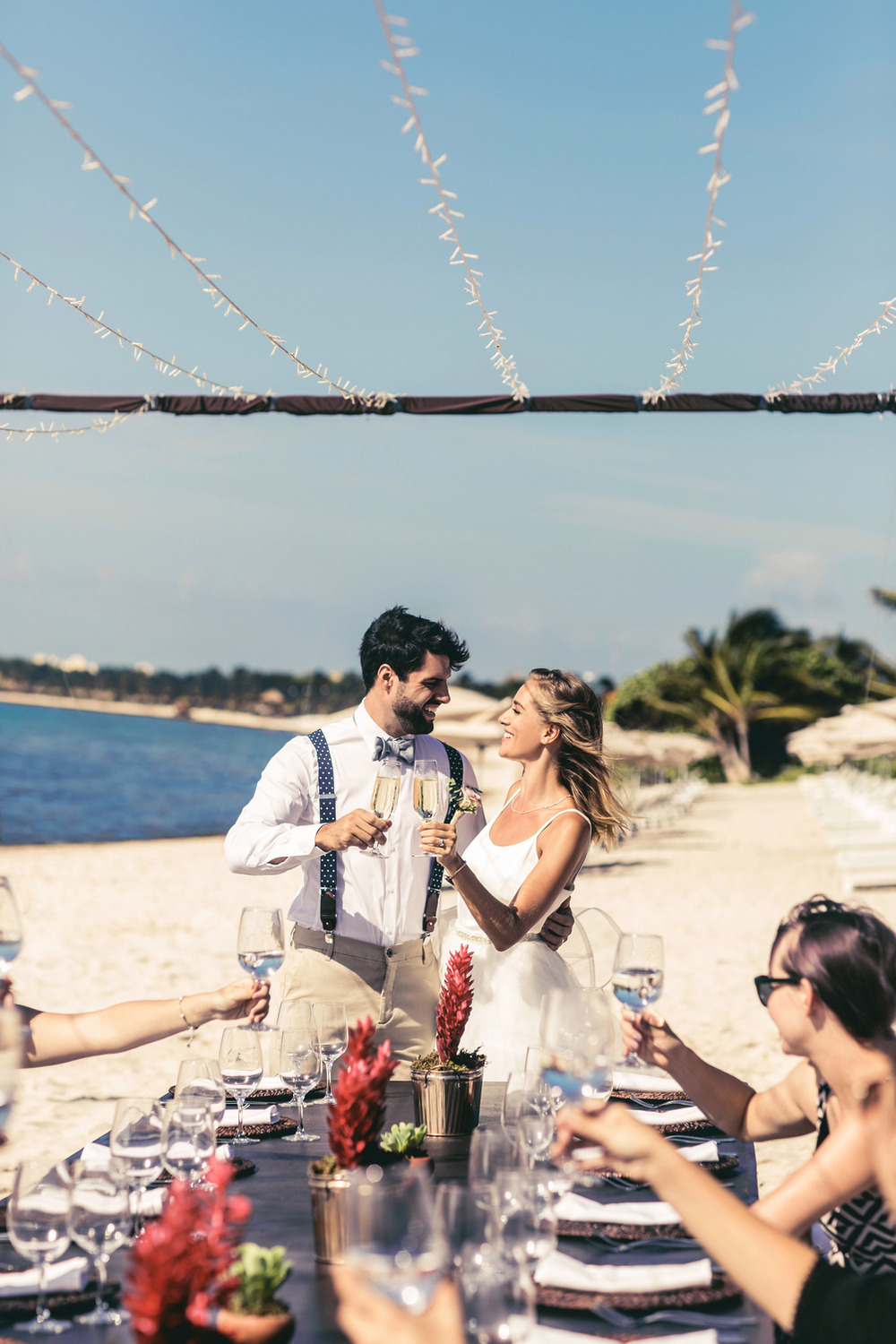 A couple raises their glasses at a beachside table during a destination wedding, while joyful wedding music floats through the sunny sky.