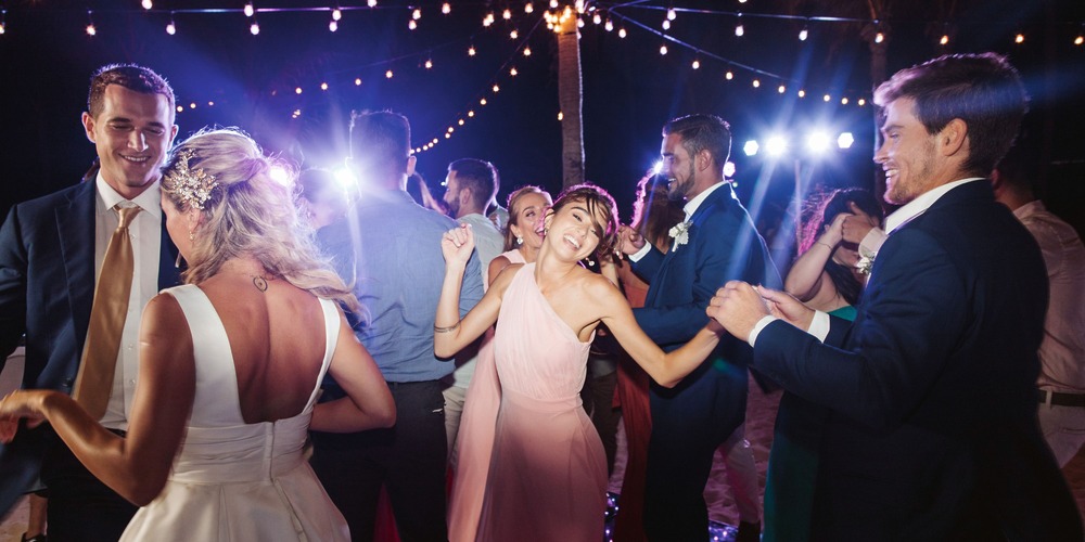Elegantly dressed guests dance to lively wedding music beneath glowing string lights at an outdoor destination wedding celebration under the night sky.