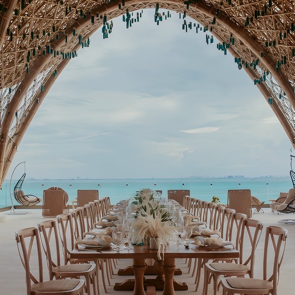 Long banquet table set for a destination wedding under a bamboo canopy, with views of the ocean.