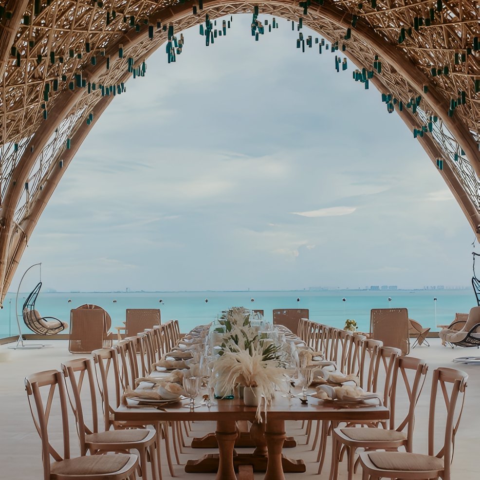 Long banquet table set for a destination wedding under a bamboo canopy, with views of the ocean.