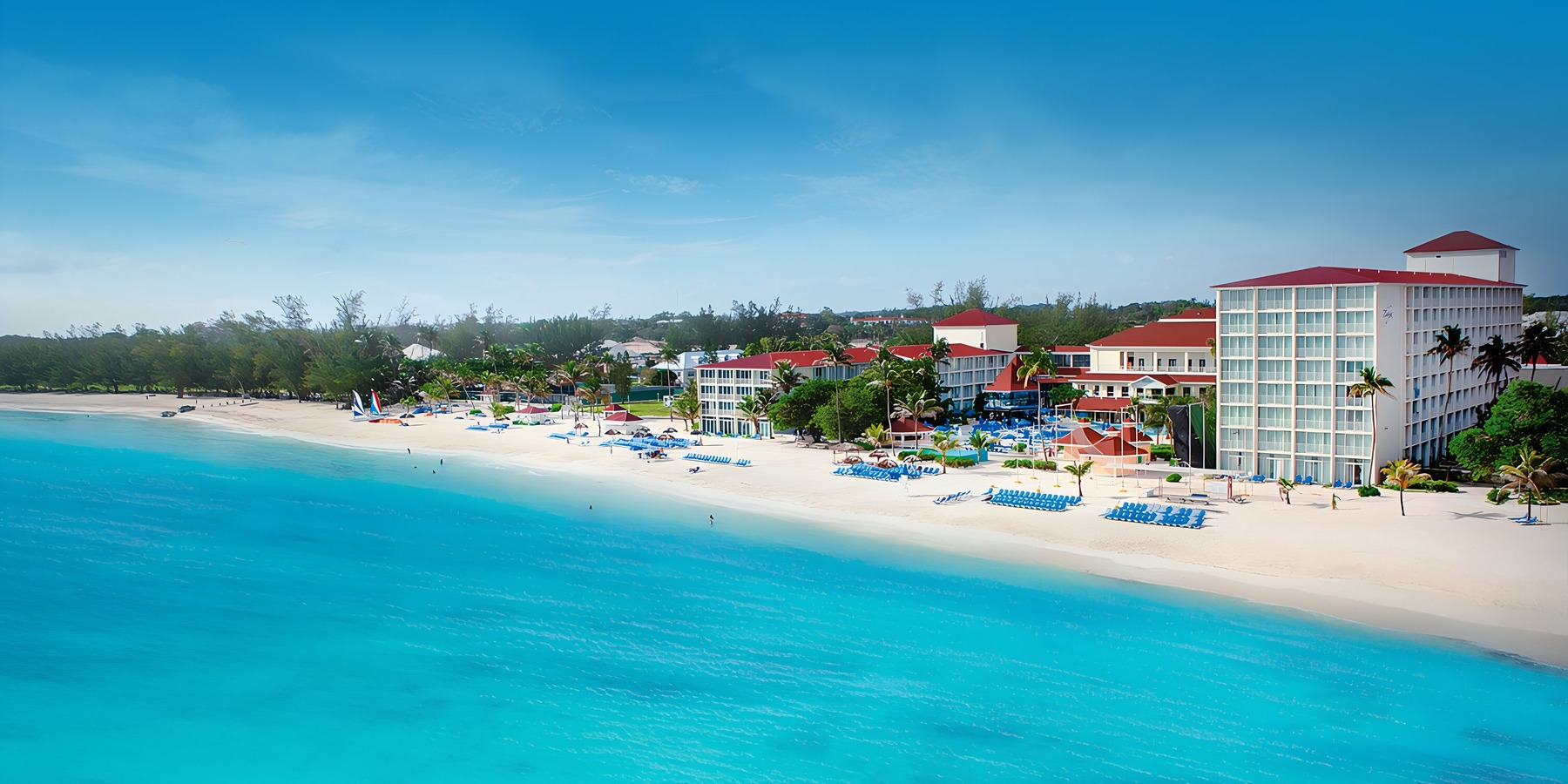 Aerial view of a tropical beach resort with white buildings, palm trees, and people relaxing by the turquoise sea.