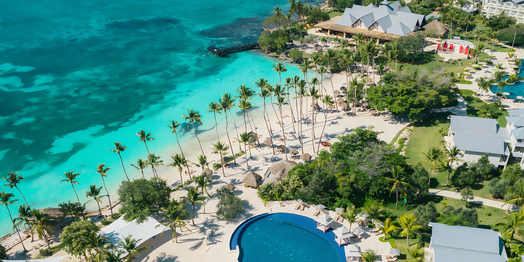 Aerial view of a tropical resort with a swimming pool, palm trees, and a clear blue ocean.