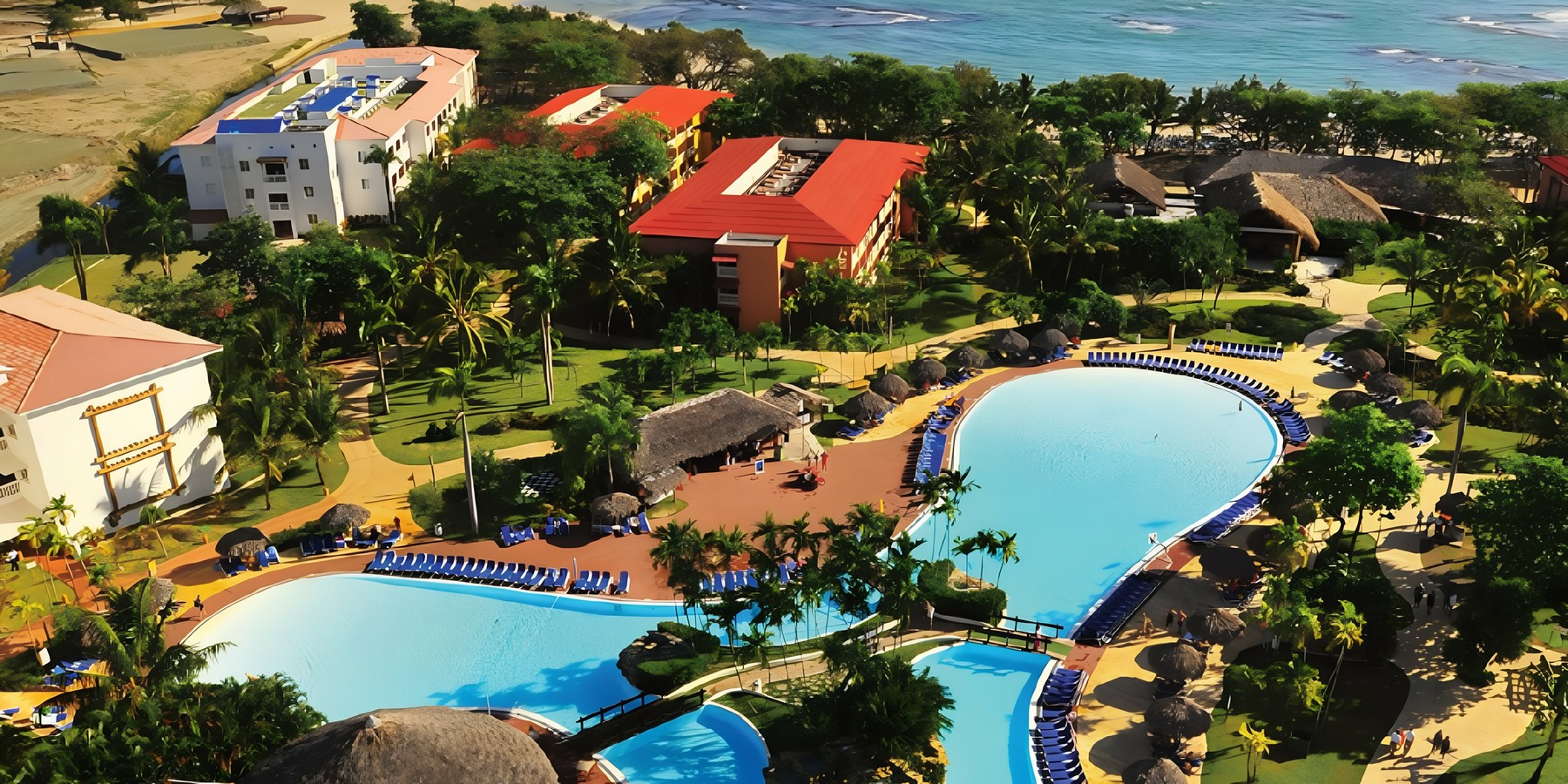 Aerial view of a coastal resort with large swimming pools, palm trees, and adjacent beach.