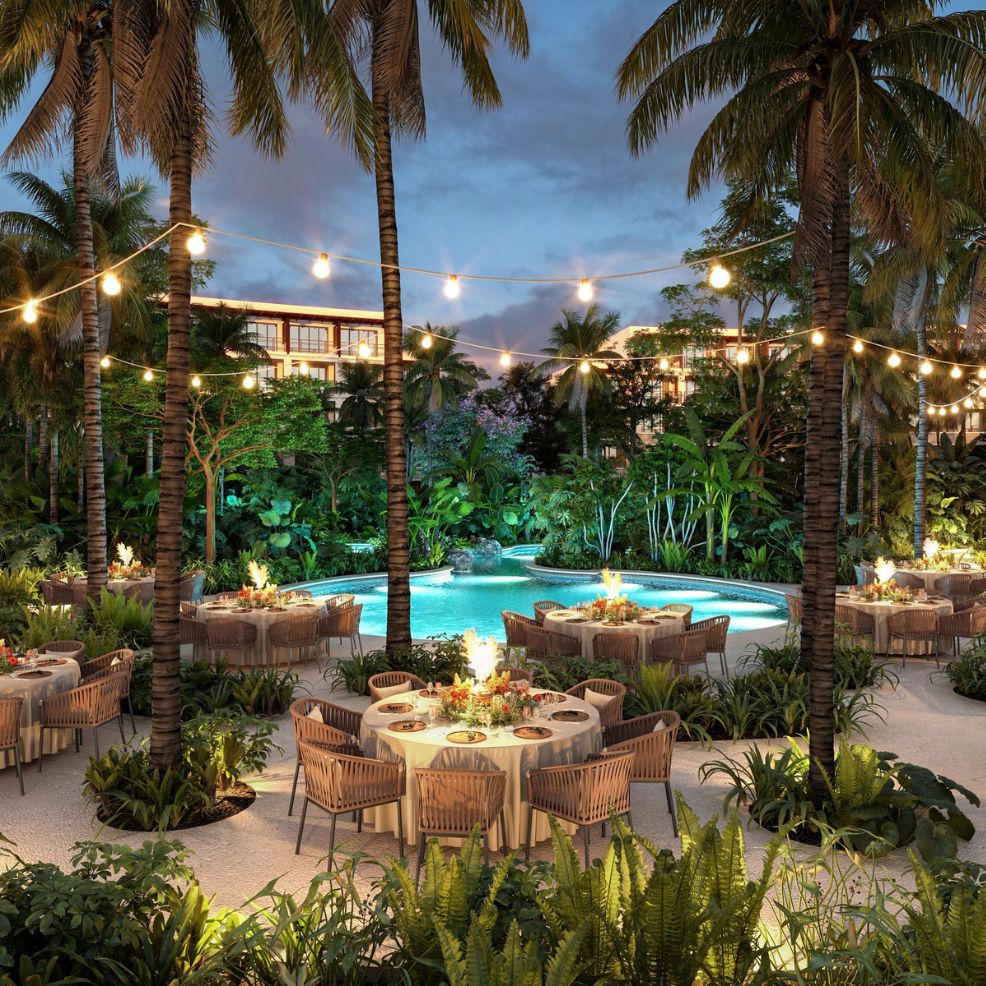 Outdoor dining tables for a destination wedding surround a shimmering pool, illuminated by string lights and framed by swaying palm trees at dusk.
