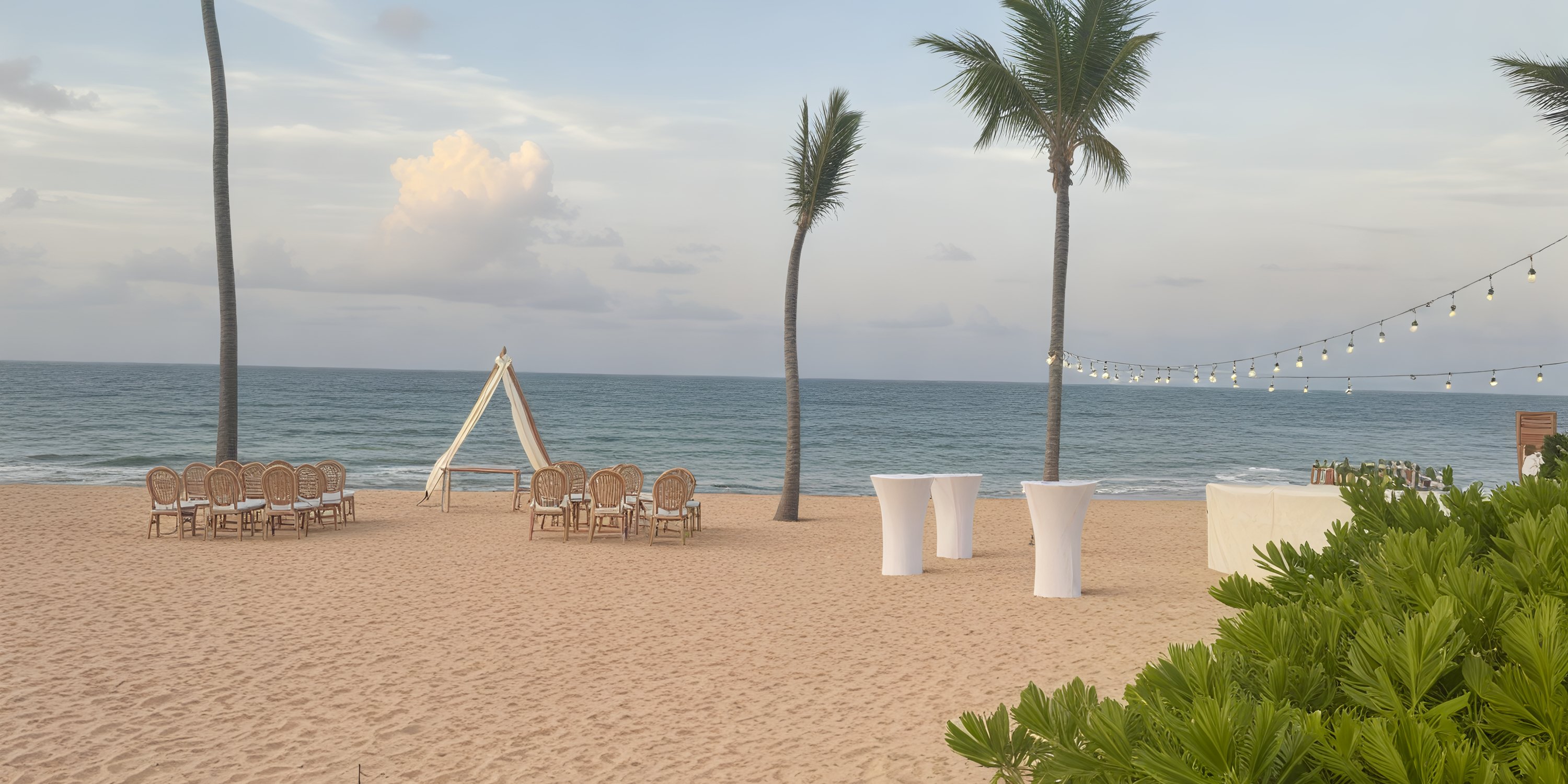 Secrets Tides destination wedding setup: chairs and tables elegantly arranged beneath enchanting string lights by the ocean, creating a romantic scene under a cloudy sky.