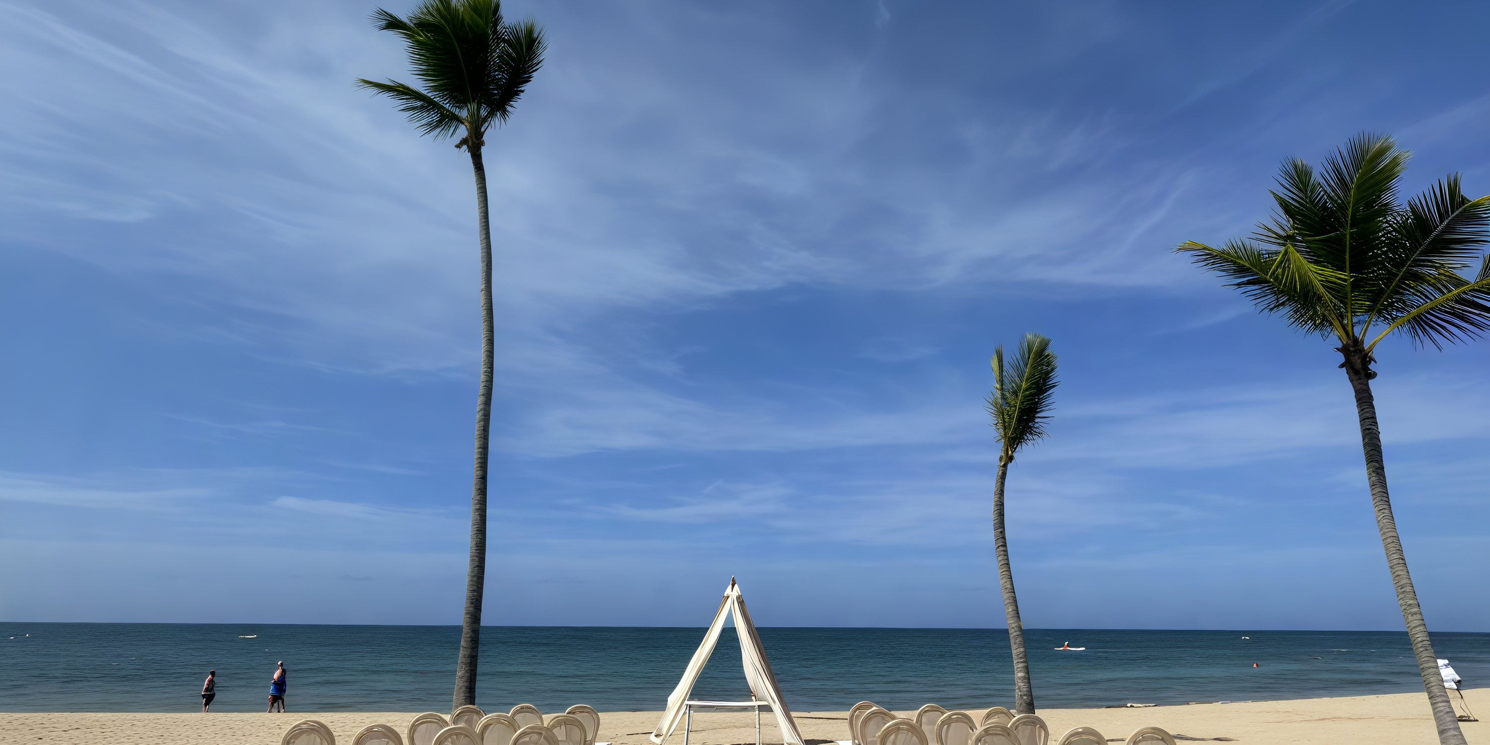 Two tall palms frame the wedding arch at Secrets Tides, set on a sandy beach beneath a brilliant blue sky—the perfect setting for a destination wedding.