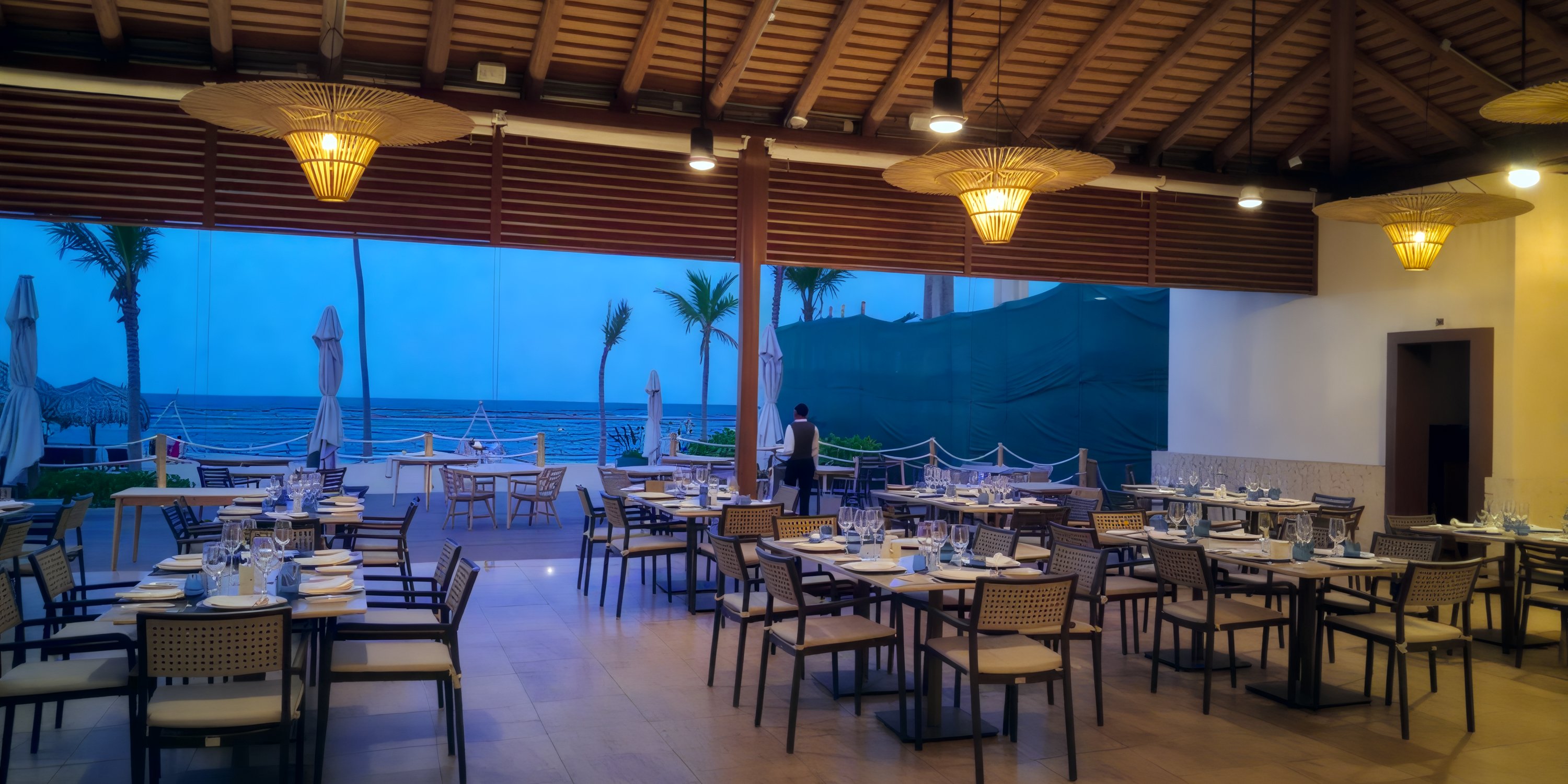 Secrets Tides: Open-air dining perfect for a destination wedding, set tables, wicker chairs, dusk beach view.