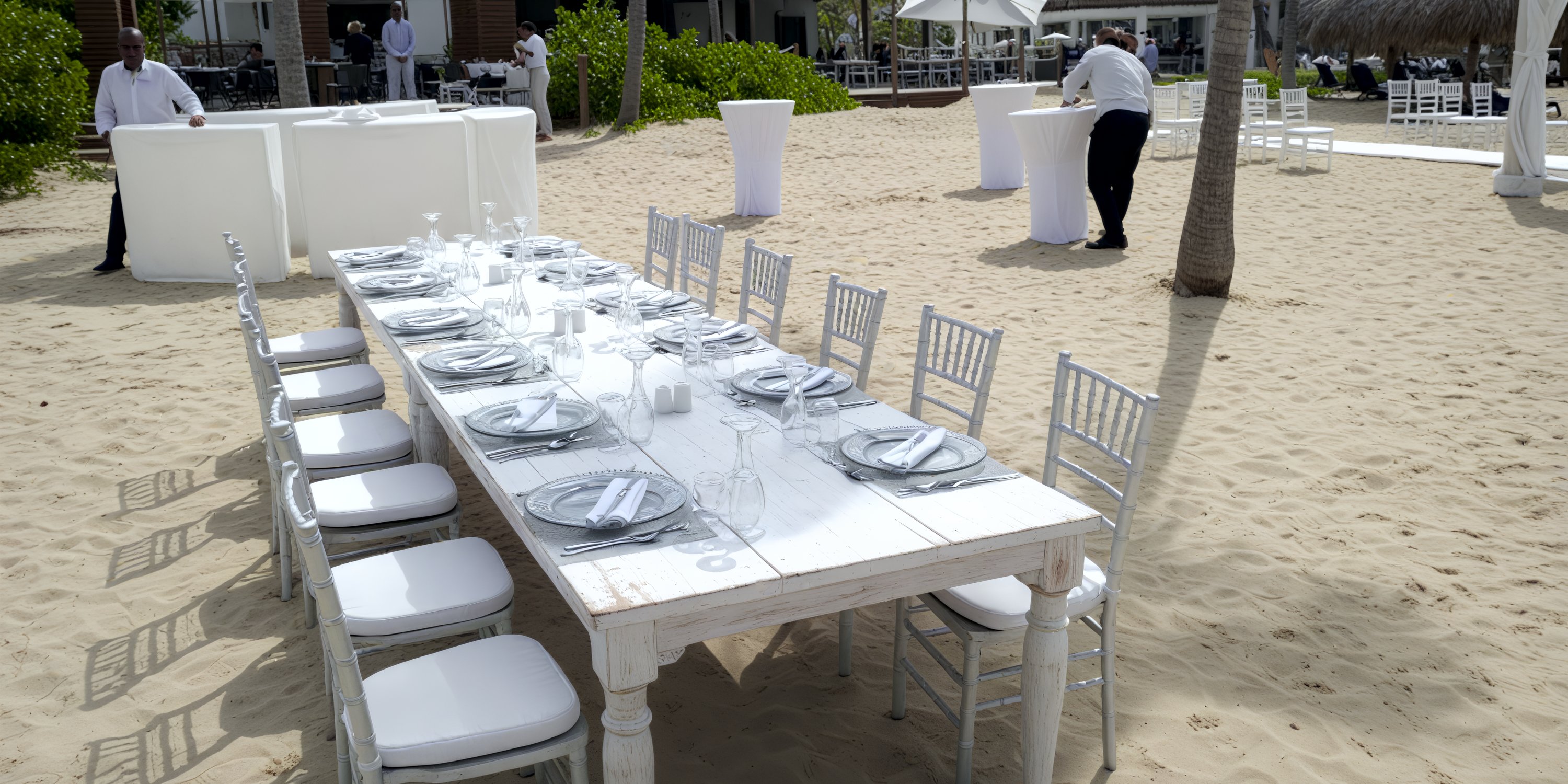 Long white table set for a destination wedding meal on the beach, with chairs and staff nearby.