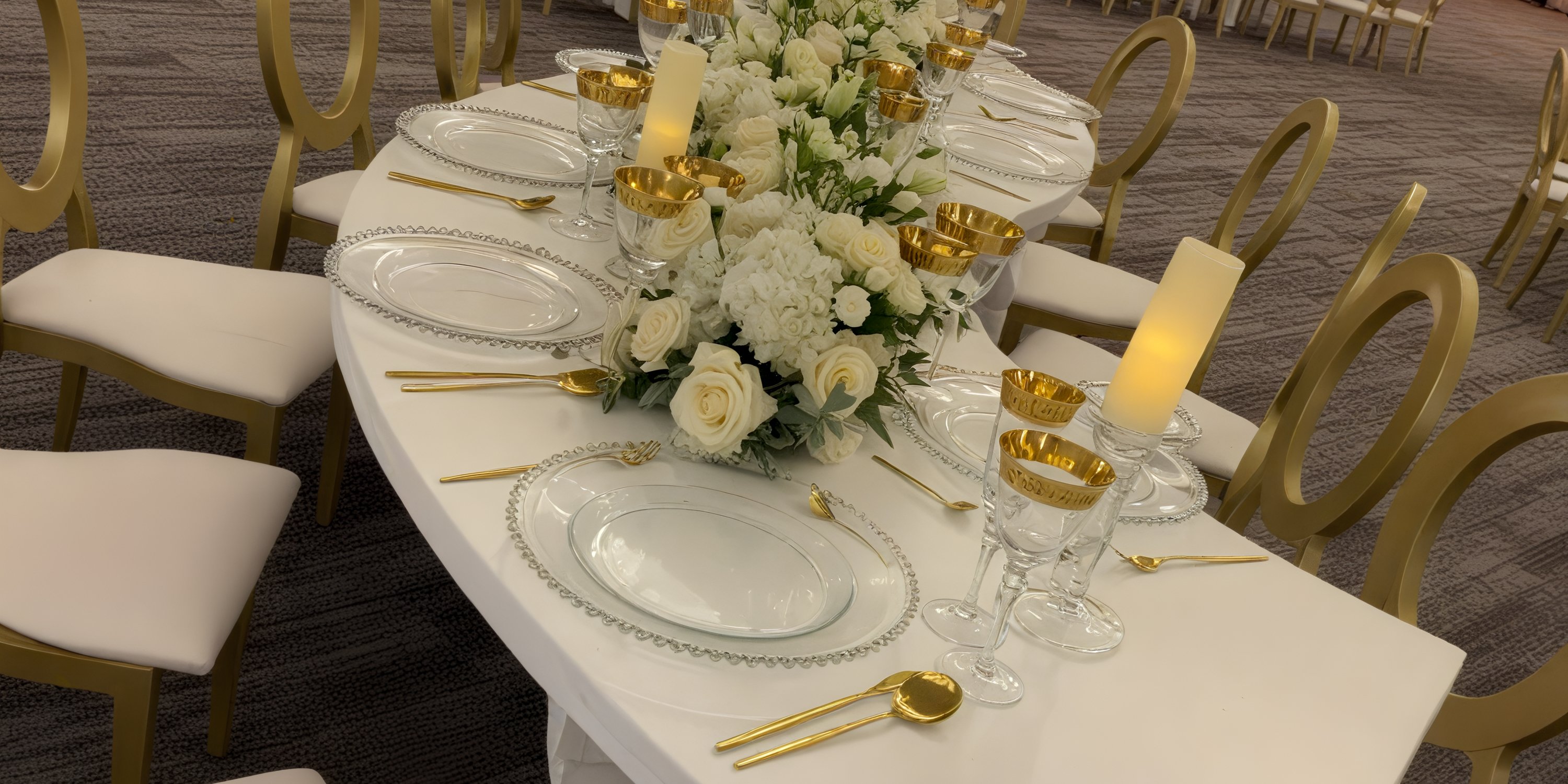 Destination wedding table setting with white flowers, gold utensils, and clear plates on white linens.