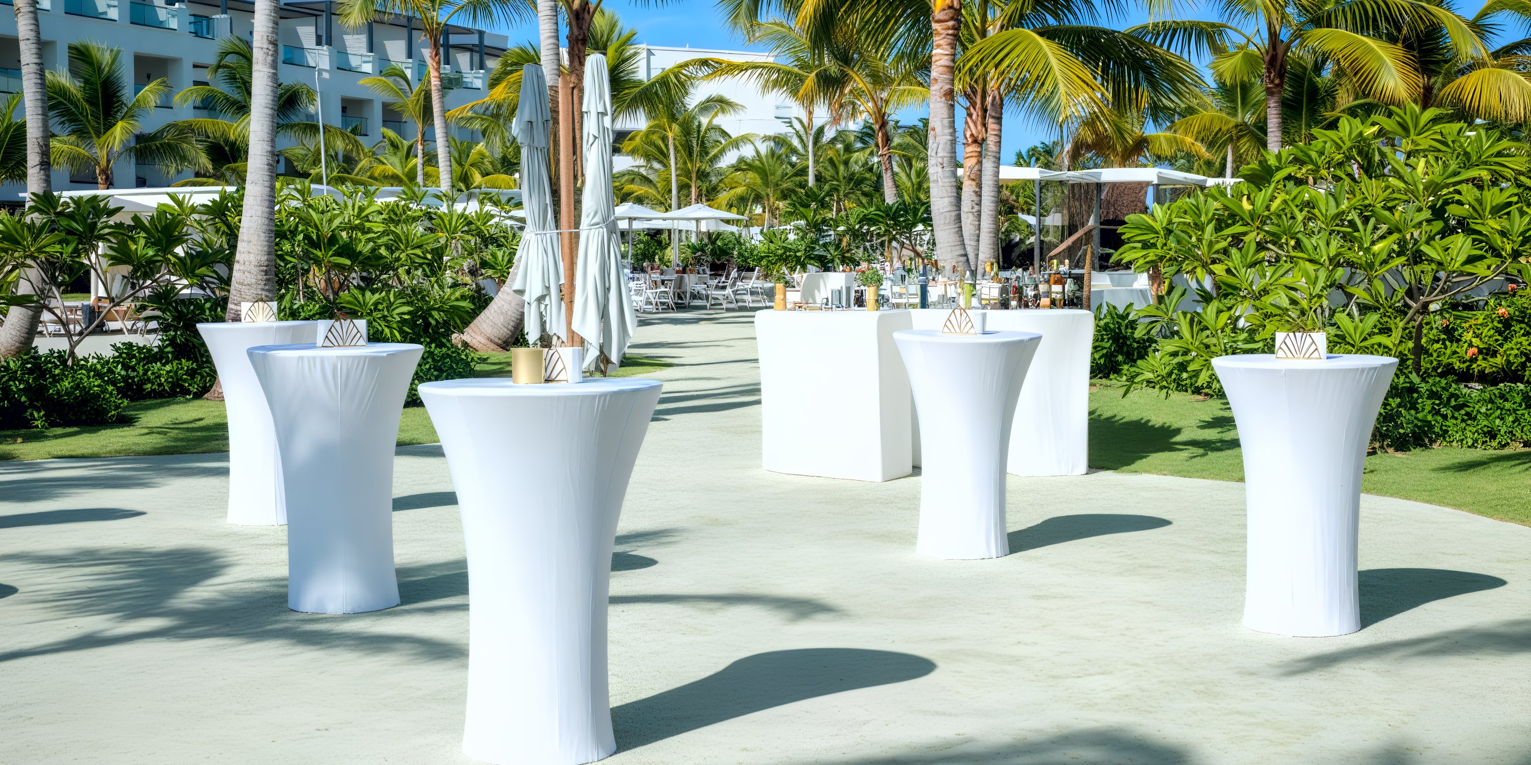Destination wedding event setup with white cocktail tables and palm trees in a lush resort garden.