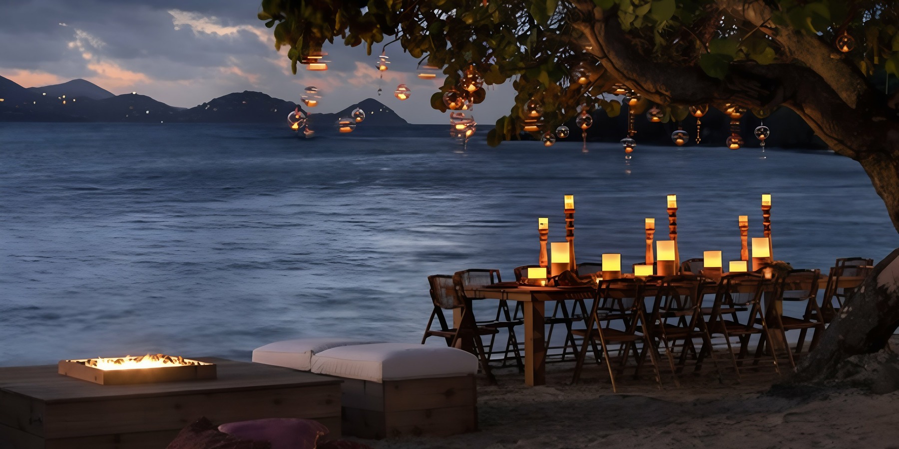 Evening beachside dining setup with lit candles on tables, hanging lanterns from a tree, and a view of the sea and distant mountains.