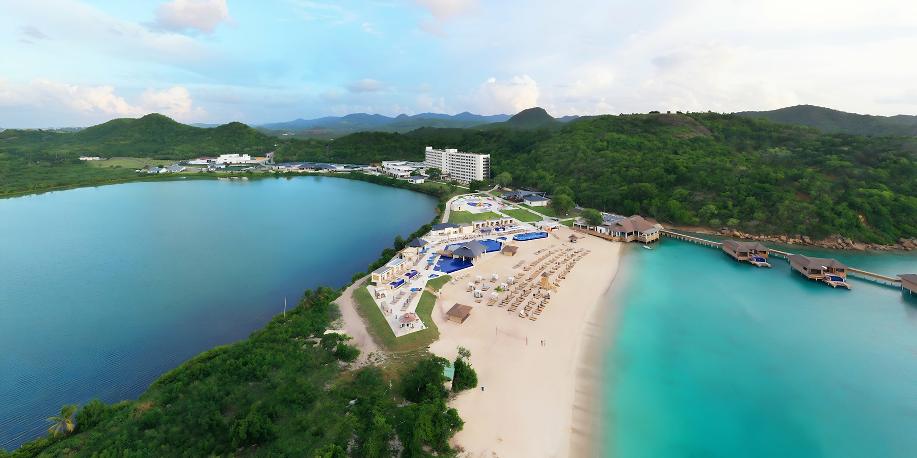 Aerial view of a tropical resort with a hotel, sandy beach, and a calm bay bordered by lush green hills.
