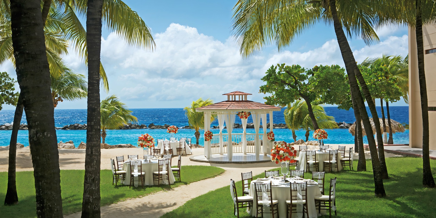Outdoor wedding setup on a beach with a white gazebo, chairs arranged for guests, and palm trees overlooking the ocean.