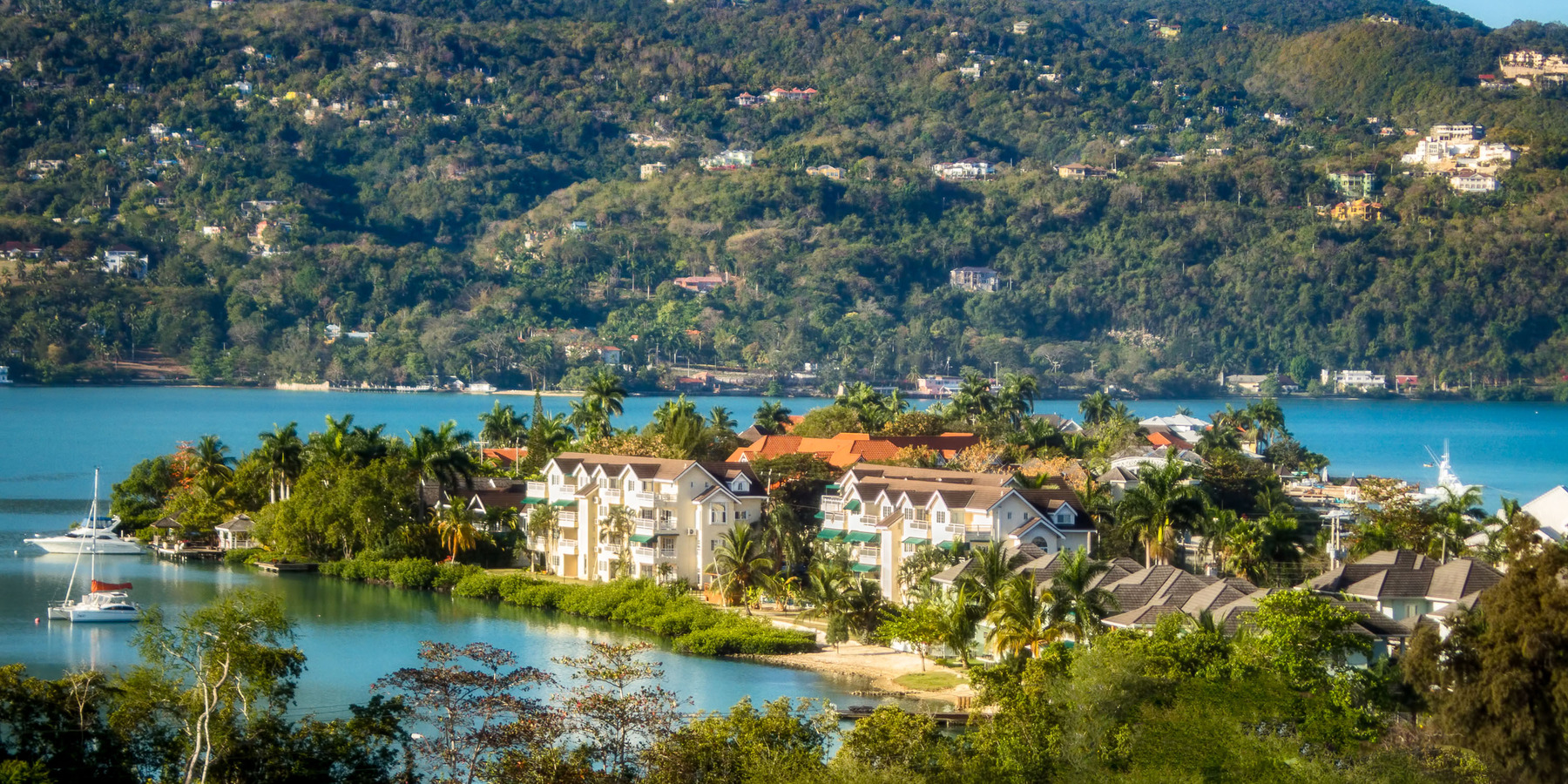 Lush green hillside overlooking a tropical coastal town with clusters of white buildings and boats moored in the blue water.