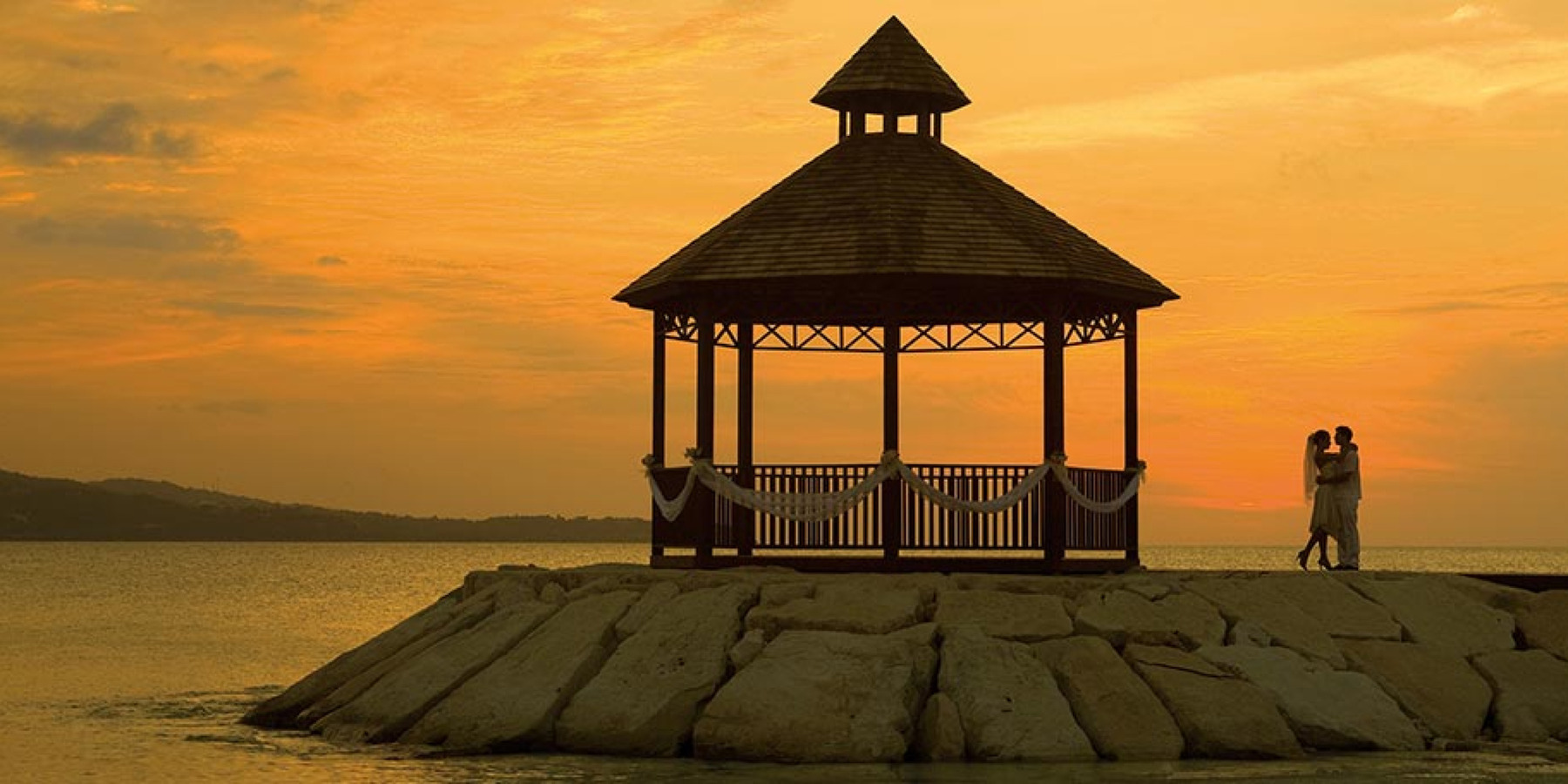 A couple embraces beside a gazebo on a rocky shoreline against a sunset sky.