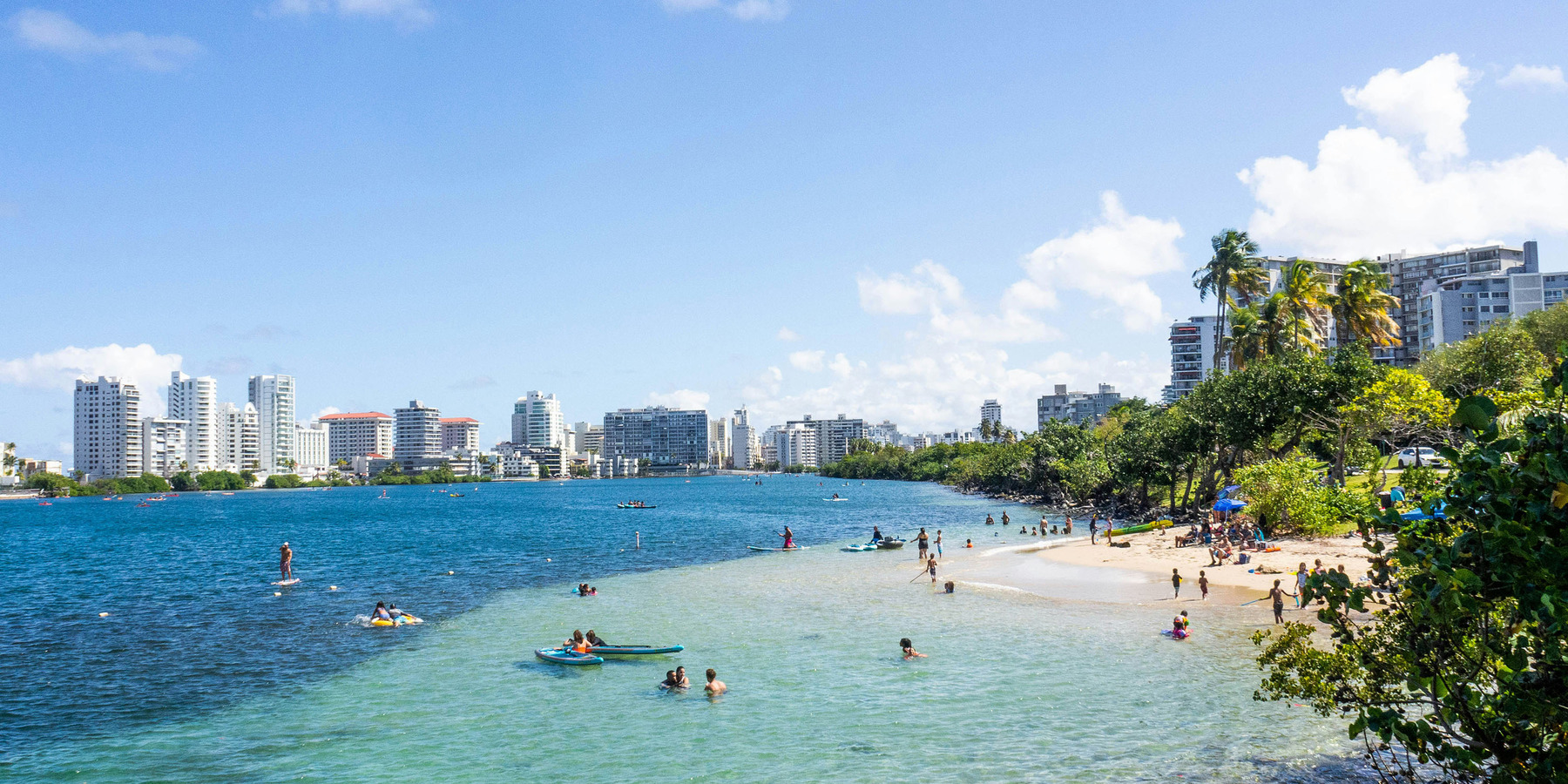 Urban beach with people enjoying water activities on a sunny day, with city skyline in the background.