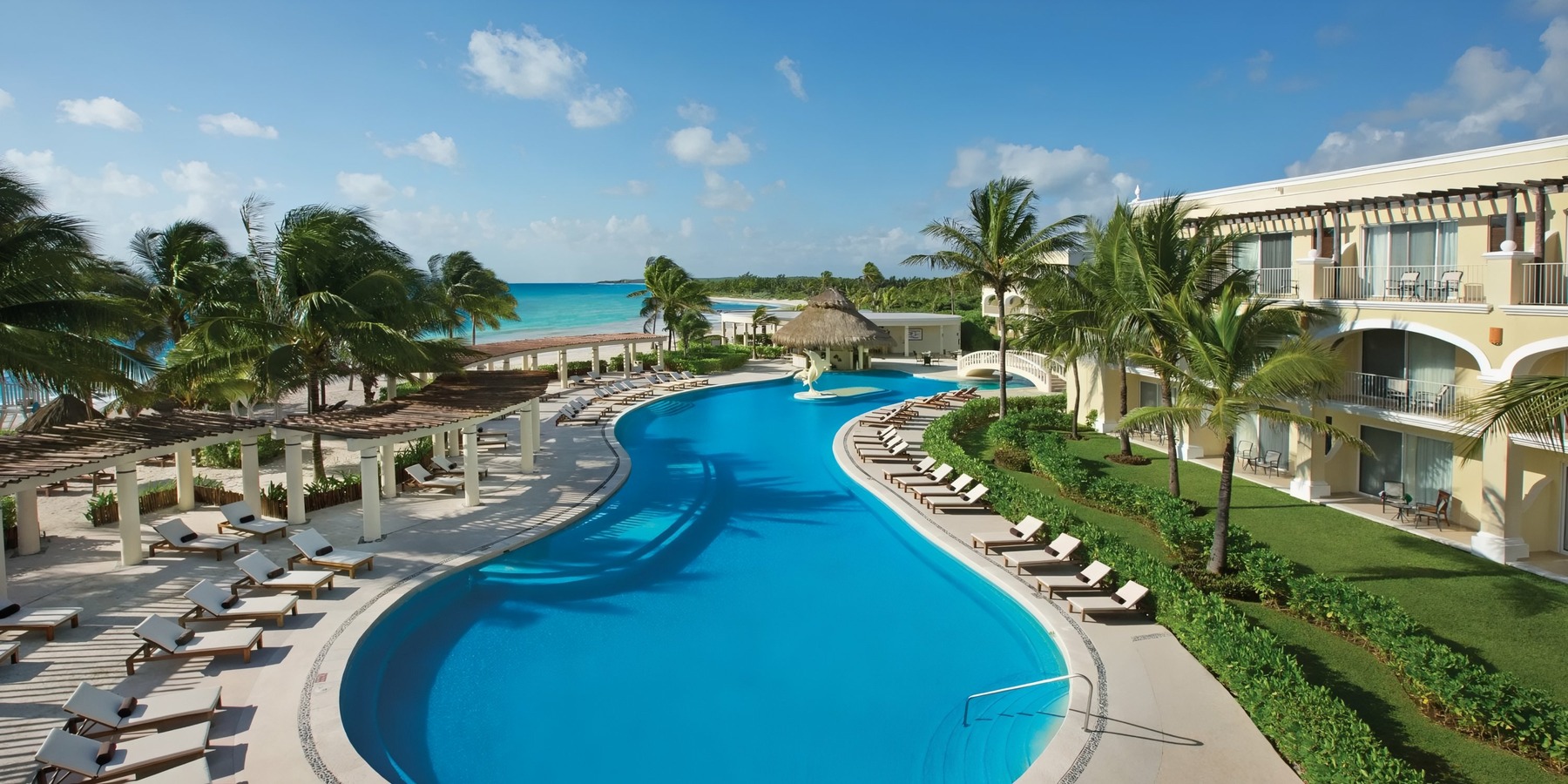 Luxury beachfront resort with a large curving swimming pool surrounded by lounge chairs, flanked by palm trees, and overlooking a calm turquoise sea.