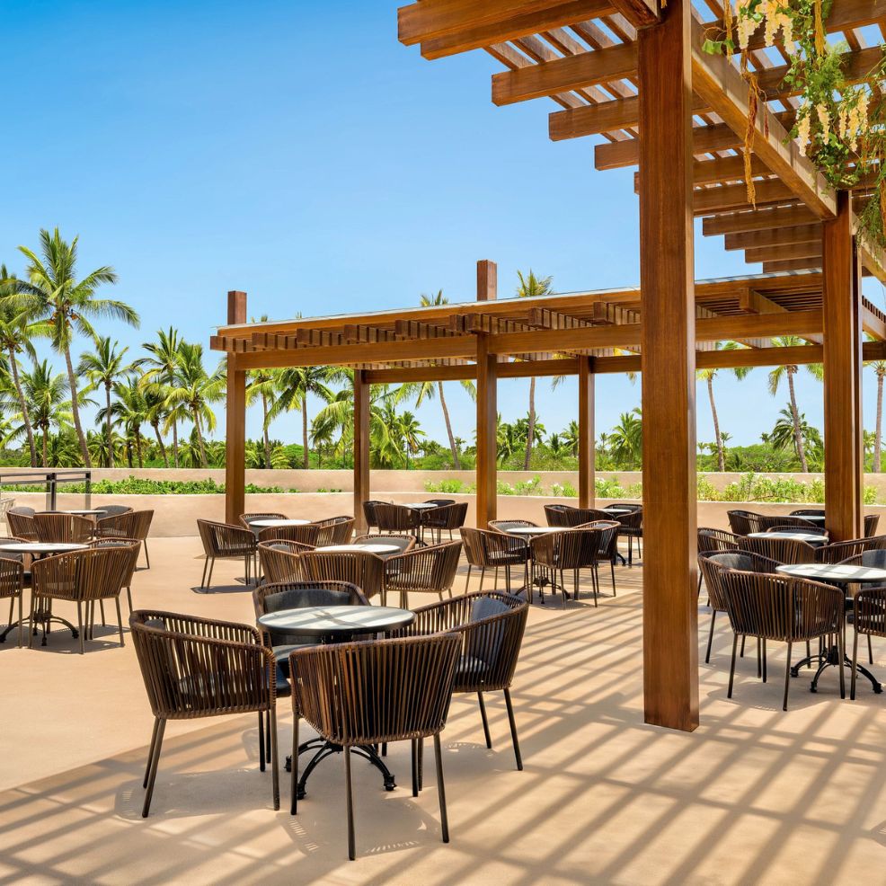 Outdoor patio set for a destination wedding, empty tables and chairs under pergolas, palm trees behind.