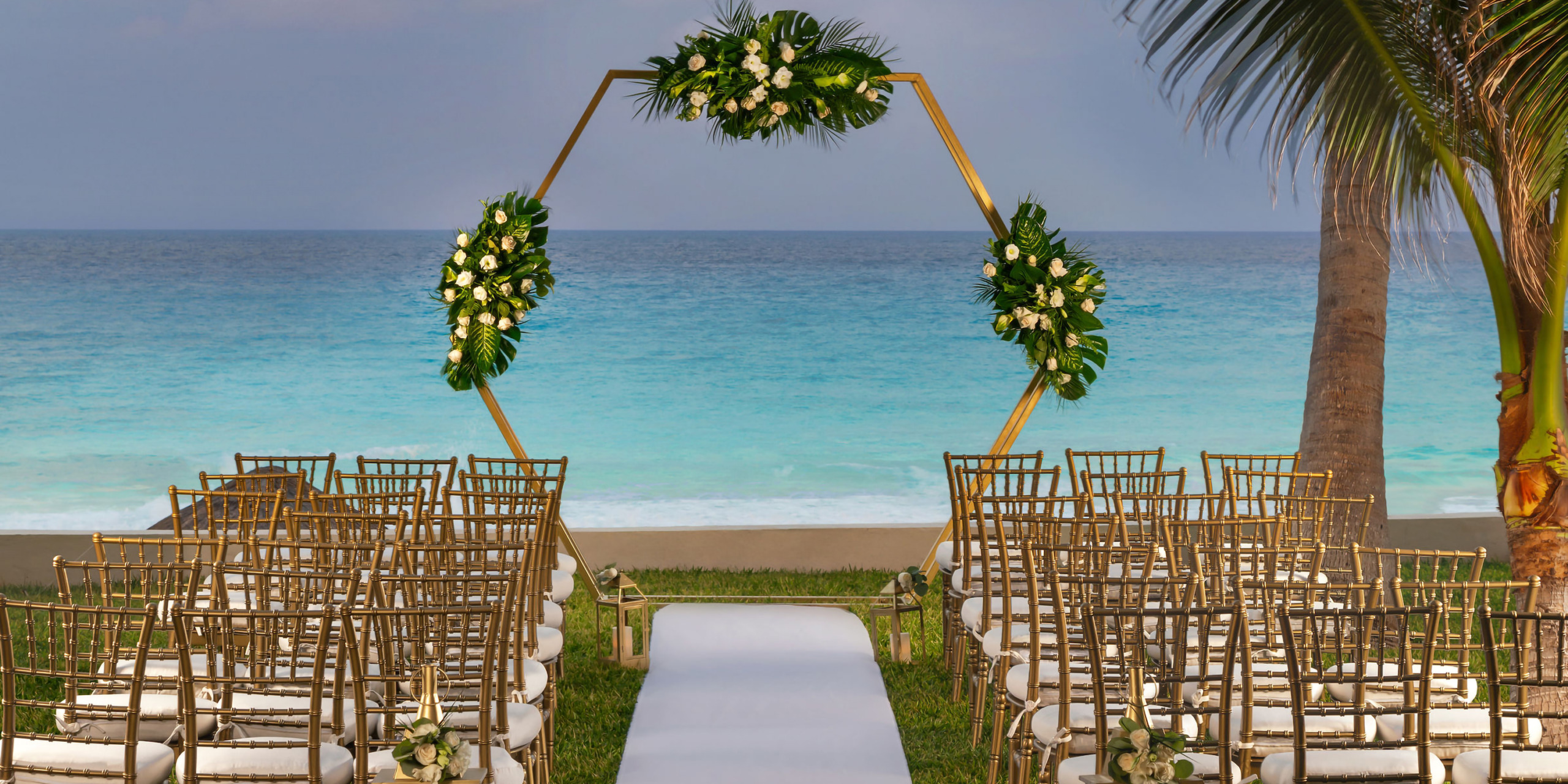Outdoor wedding setup at the Hilton Cancun Mar Caribe with rows of gold chairs, a white aisle, and a floral arch, overlooking the ocean.