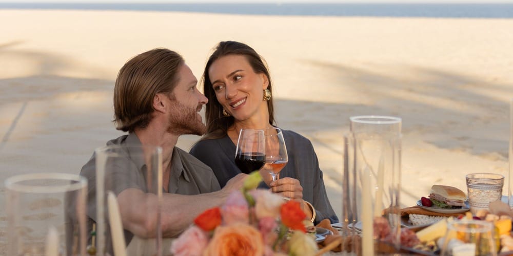 Newlyweds celebrate their destination wedding, sipping drinks and gazing at each other by the beach.