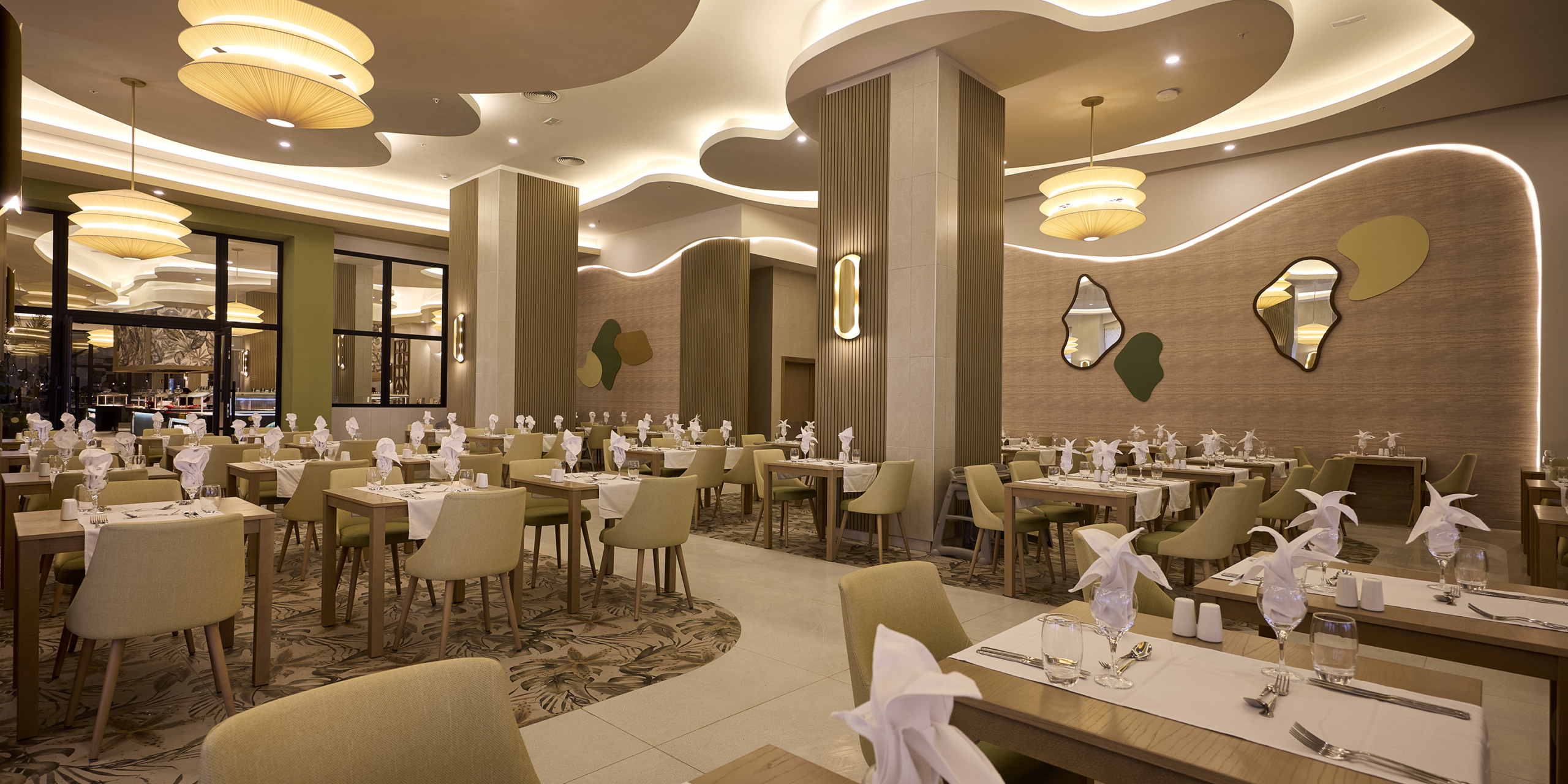 A modern restaurant with neatly arranged tables, green and beige chairs, folded napkins, and abstract wall decorations. Soft lighting from pendant lamps creates a warm ambiance at RIU Palace Aquarelle.