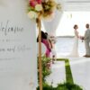At the picturesque Sandals South Coast, a couple stands at a captivating outdoor wedding ceremony by the water. In the foreground, a beautifully adorned welcome sign enhances the warm and inviting atmosphere of this unforgettable day.