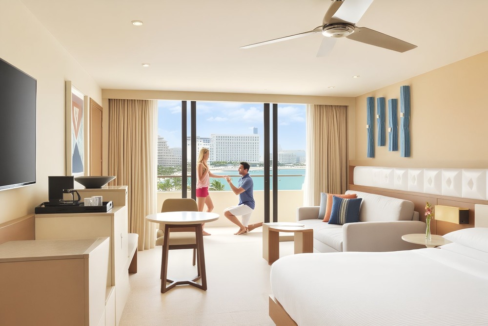 A man proposes to a woman in a sunlit hotel room, drawing inspiration from heartfelt wedding vow examples—setting the stage for their unforgettable destination wedding.