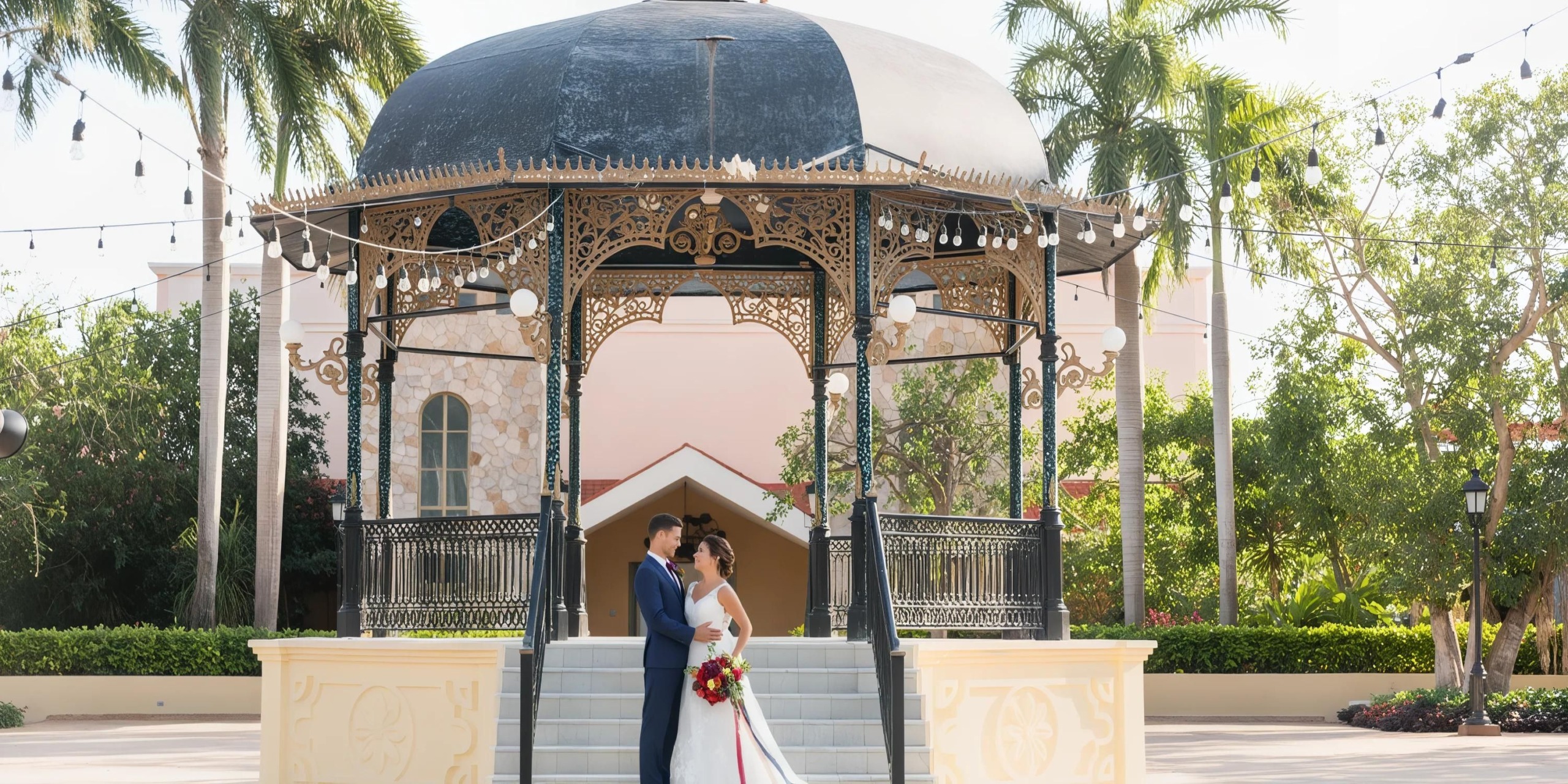 A couple in wedding attire stands facing each other under an ornate, open-air pavilion with steps, surrounded by trees and string lights.