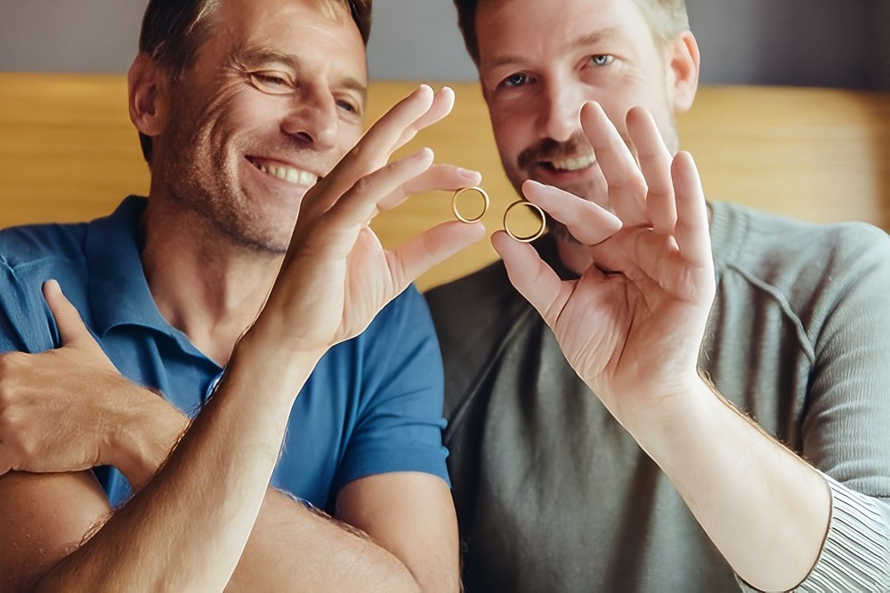 Two men smiling and proudly holding up gold rings together after sharing examples of wedding vows at their destination wedding.