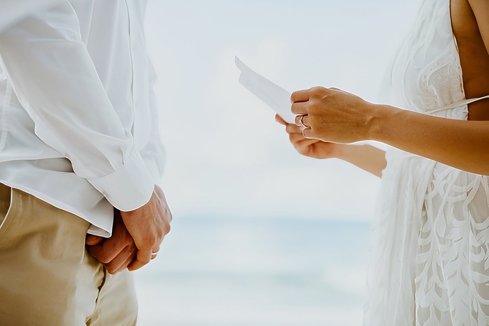 At a beachside destination wedding, a person in a white dress reads examples of wedding vows to their partner during the ceremony.