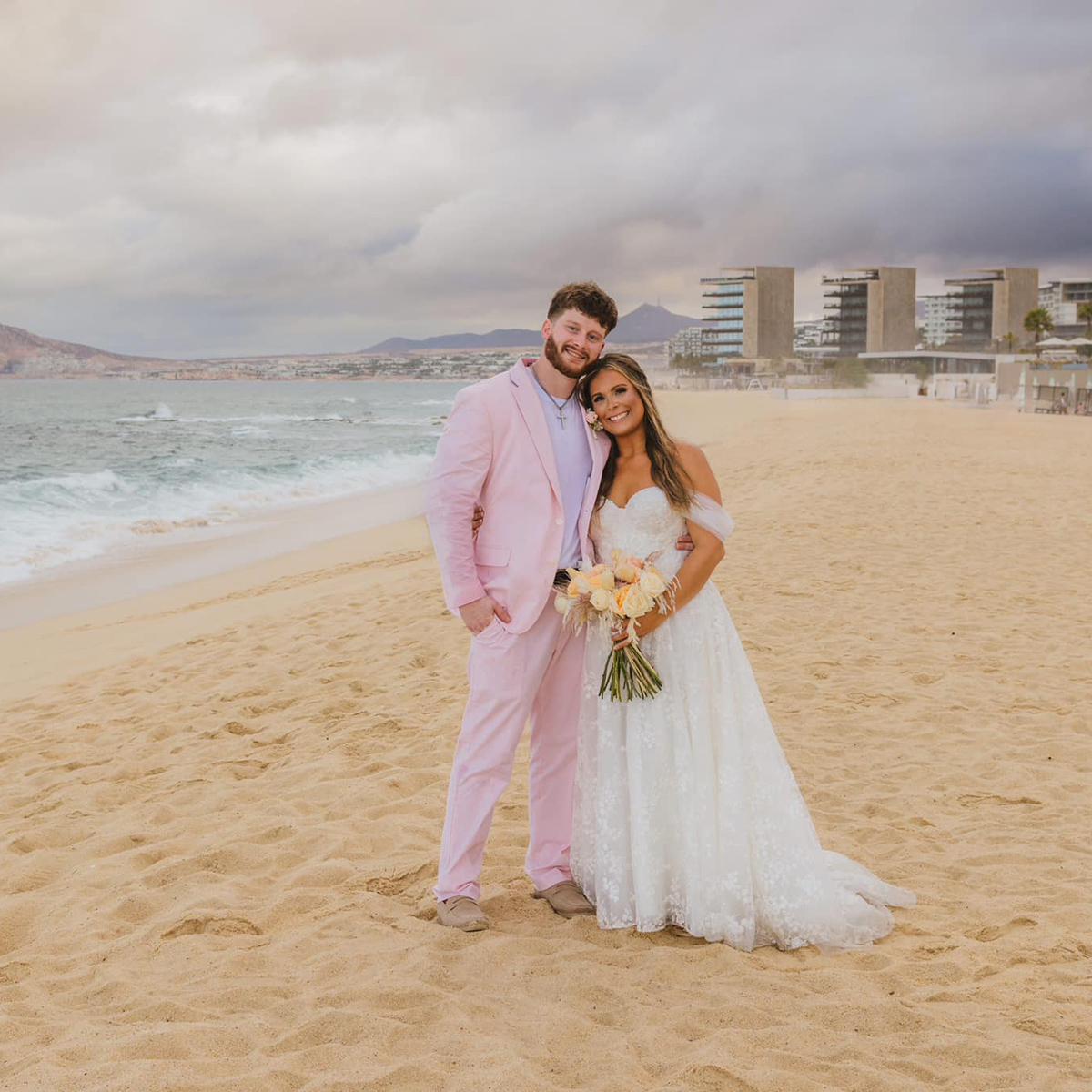 A couple stands on a beach with buildings in the background. The man wears a pink suit, and the woman, wearing the "Anna Grace Hall" white wedding dress and holding a bouquet of flowers, stands beside him.