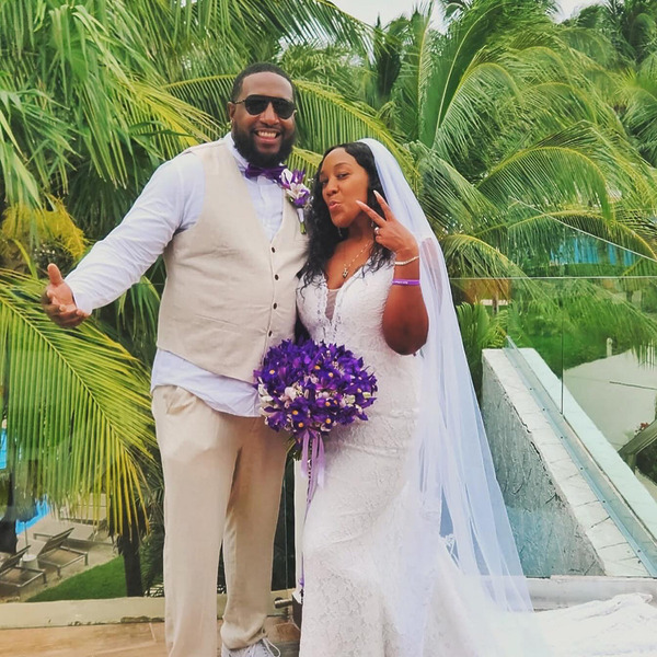 A newlywed couple posing outdoors in front of palm trees, with the groom dressed in a beige suit and the bride in a Cherise Dawkins white wedding dress, holding a purple bouquet.