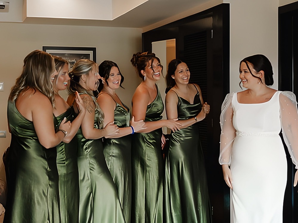 Six bridesmaids, dressed in green summer wedding guest dresses, react with delight to the bride as she appears in her white gown indoors at the destination wedding.