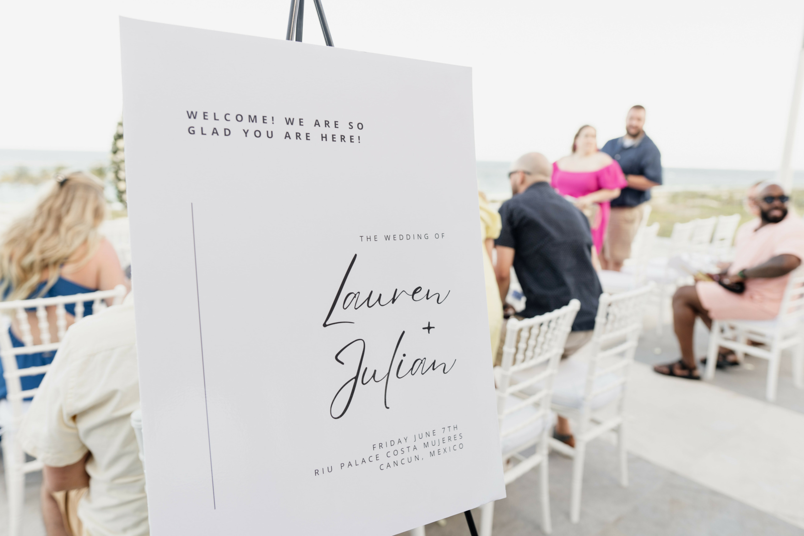 A wedding sign reads "Welcome! We are so glad you are here! The wedding of Lauren and Julian", with guests seated in the background.