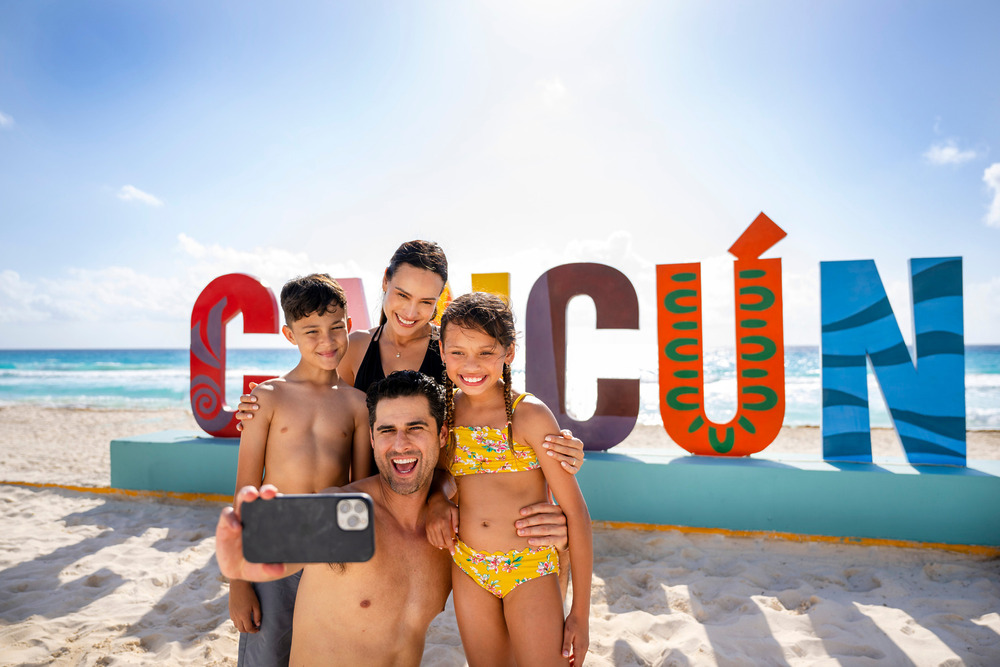 A family poses for a beach selfie in front of the vibrant "CANCUN" letters, chatting about the cost of a Riviera Maya destination wedding.