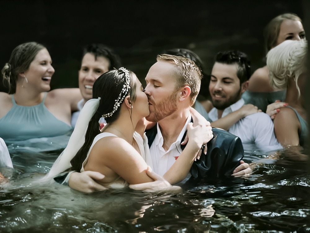 The bride and groom share a kiss in the water, creating an iconic destination wedding photo, surrounded by their smiling friends.