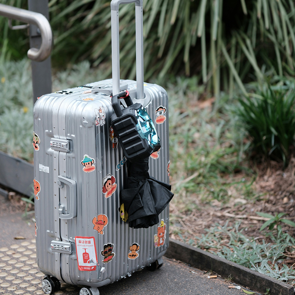 A silver suitcase adorned with colorful stickers stands on a paved path by greenery, with a black and blue umbrella attached to its handle.