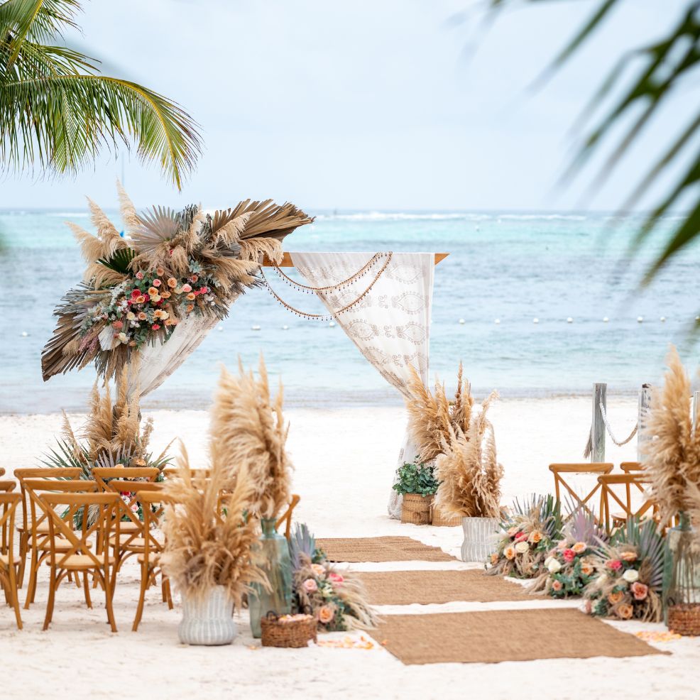 Destination wedding setup on the beach featuring pampas grass, floral decor, and wooden chairs by the ocean.