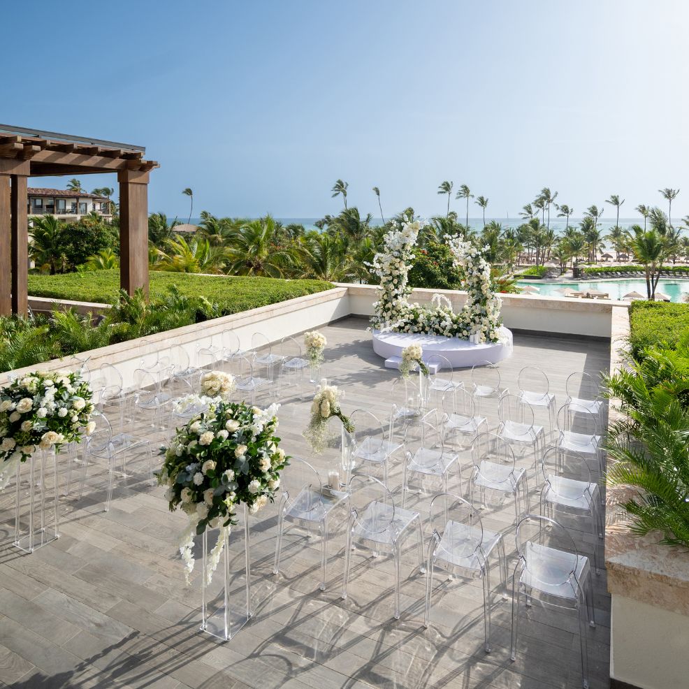 Destination wedding ceremony arrangement featuring clear chairs, white flowers, and a lush tropical backdrop.