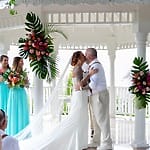 A couple kisses at their destination wedding in Jamaica beneath a white gazebo, guests watching.